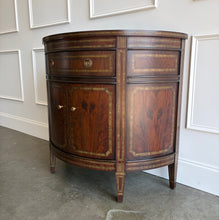Load image into Gallery viewer, The Demilune Cabinet by Ambella Home combines graceful curves with richly figured wood veneer and classical detailing. Antiqued gold accents outline the drawer, doors, architectural panels, and tapered legs, emphasizing the cabinet’s rounded silhouette. A spacious upper drawer with decorative ring pulls provides convenient storage for smaller essentials, while two curved doors open to reveal an interior shelf for larger items. Good Bones Furniture