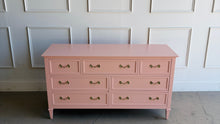Load image into Gallery viewer, This vintage 7 drawer dresser by Drexel Furniture has been freshly lacquered in Benjamin Moore Mellow Coral, a warm blush-pink finish that gives the piece a soft, elevated look without losing its classic character. The clean rectangular case, framed drawer fronts, tapered legs, and original polished brass hardware create a tailored silhouette with subtle traditional detail. Good Bones Furniture