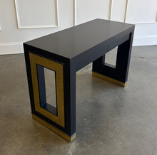 Load image into Gallery viewer, The Ashleigh Console by Theodore Alexander pairs a streamlined silhouette with dramatic decorative details. Professionally lacquered in black, it features substantial rectangular supports accented by textured gold panels, inset antiqued mirror, and brass-capped feet. Two discreet drawers provide concealed storage. Good Bones Furniture
