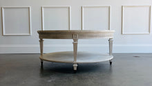 Load image into Gallery viewer, The Lucienne Cocktail Table by Theodore Alexander combines classical ornament with a light, graceful silhouette. Crafted from Khaya wood and richly figured Khaya swirl veneer, its round top rests above a fluted frieze accented with carved leaf motifs. Slender fluted legs connect the upper surface to a wide circular lower shelf. Washed Blanchette finish. Good Bones Furniture