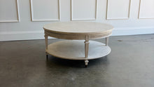 Load image into Gallery viewer, The Lucienne Cocktail Table by Theodore Alexander combines classical ornament with a light, graceful silhouette. Crafted from Khaya wood and richly figured Khaya swirl veneer, its round top rests above a fluted frieze accented with carved leaf motifs. Slender fluted legs connect the upper surface to a wide circular lower shelf. Washed Blanchette finish. Good Bones Furniture