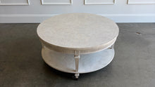 Load image into Gallery viewer, The Lucienne Cocktail Table by Theodore Alexander combines classical ornament with a light, graceful silhouette. Crafted from Khaya wood and richly figured Khaya swirl veneer, its round top rests above a fluted frieze accented with carved leaf motifs. Slender fluted legs connect the upper surface to a wide circular lower shelf. Washed Blanchette finish. Good Bones Furniture