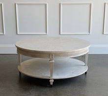 Load image into Gallery viewer, The Lucienne Cocktail Table by Theodore Alexander combines classical ornament with a light, graceful silhouette. Crafted from Khaya wood and richly figured Khaya swirl veneer, its round top rests above a fluted frieze accented with carved leaf motifs. Slender fluted legs connect the upper surface to a wide circular lower shelf. Washed Blanchette finish. Good Bones Furniture