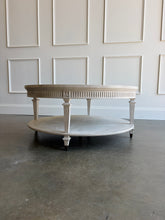 Load image into Gallery viewer, The Lucienne Cocktail Table by Theodore Alexander combines classical ornament with a light, graceful silhouette. Crafted from Khaya wood and richly figured Khaya swirl veneer, its round top rests above a fluted frieze accented with carved leaf motifs. Slender fluted legs connect the upper surface to a wide circular lower shelf. Washed Blanchette finish. Good Bones Furniture