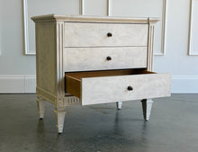 Load image into Gallery viewer, The Valette Nightstand by Theodore Alexander combines generous bedside storage with graceful architectural detail. Crafted from Khaya wood and richly figured Khaya swirl veneer, its three spacious drawers are accented with sculptural floral metal pulls. Fluted corner posts frame the case, while carved leaf motifs, a fluted apron, and tapered legs introduce classical character without overwhelming the clean silhouette. Good Bones Furniture