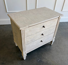 Load image into Gallery viewer, The Valette Nightstand by Theodore Alexander combines generous bedside storage with graceful architectural detail. Crafted from Khaya wood and richly figured Khaya swirl veneer, its three spacious drawers are accented with sculptural floral metal pulls. Fluted corner posts frame the case, while carved leaf motifs, a fluted apron, and tapered legs introduce classical character without overwhelming the clean silhouette. Good Bones Furniture