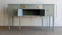 Load image into Gallery viewer, This vintage sideboard pairs a historic Hepplewhite-inspired silhouette with an unexpected modern color. Its gently bowed front, symmetrical layout, slender tapered legs, and balanced proportions reflect classic Federal design. Professionally lacquered in Sherwin-Williams Unusual Gray with new solid brass oval bail pulls with ornate neoclassical backplates. Good Bones Furniture