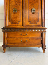 Load image into Gallery viewer, This vintage Mediterranean armoire has bold 1970s Spanish Revival styling with carved frieze detail, stepped door panels, burl veneer accents, and original brass ring hardware. The warm walnut-toned finish highlights the depth of the veneer, while the interior offers generous concealed storage with built-in drawers. Good Bones Furniture