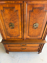 Load image into Gallery viewer, This vintage Mediterranean armoire has bold 1970s Spanish Revival styling with carved frieze detail, stepped door panels, burl veneer accents, and original brass ring hardware. The warm walnut-toned finish highlights the depth of the veneer, while the interior offers generous concealed storage with built-in drawers. Good Bones Furniture