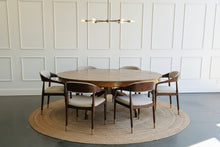 Load image into Gallery viewer, The Williamsburg Dining Table combines dramatic wood grain with a graceful, sculptural base. Crafted from mahogany solids, its expansive round top is available in your choice of richly figured burled ash or cerejeira veneer. Carefully arranged veneer panels create movement across the tabletop, while the substantial veneered apron adds visual depth. Four sweeping legs extend from the central pedestal and terminate in brass-capped feet. Good Bones Furniture