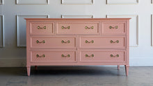 Load image into Gallery viewer, This vintage 7 drawer dresser by Drexel Furniture has been freshly lacquered in Benjamin Moore Mellow Coral, a warm blush-pink finish that gives the piece a soft, elevated look without losing its classic character. The clean rectangular case, framed drawer fronts, tapered legs, and original polished brass hardware create a tailored silhouette with subtle traditional detail. Good Bones Furniture
