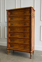 Load image into Gallery viewer, This stately Louis XVI–style chest combines generous storage with a tall, space-conscious silhouette. Crafted from mahogany solids with richly figured cerejeira veneers, it features seven spacious drawers framed by hand-applied gold accents. Fluted corner columns, paneled sides, decorative metal knobs, and tapered legs. Good Bones Furniture
