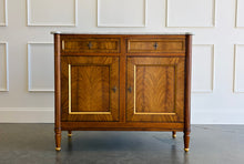 Load image into Gallery viewer, The Riley 2 Door Sideboard brings the graceful symmetry of European neoclassical design to a versatile, more compact scale. Crafted from mahogany solids with beautifully bookmatched cerejeira veneers, each door showcases a dramatic cathedral grain framed by hand-painted gold detailing. Two drawers keep smaller essentials close at hand, while the cabinet below provides generous concealed storage. Good Bones Furniture
