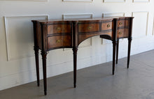 Load image into Gallery viewer, Theodore Alexander Stanhope Row Sideboard shown at an angled right view, featuring a sweeping serpentine mahogany case with flame veneers, turned uprights, fluted legs, and period brass hardware, photographed at Good Bones Furniture in Raleigh, North Carolina.
