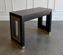 Load image into Gallery viewer, The Ashleigh Console by Theodore Alexander pairs a streamlined silhouette with dramatic decorative details. Professionally lacquered in black, it features substantial rectangular supports accented by textured gold panels, inset antiqued mirror, and brass-capped feet. Two discreet drawers provide concealed storage. Good Bones Furniture
