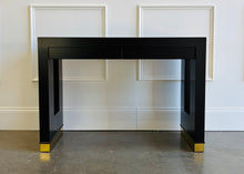 Load image into Gallery viewer, The Ashleigh Console by Theodore Alexander pairs a streamlined silhouette with dramatic decorative details. Professionally lacquered in black, it features substantial rectangular supports accented by textured gold panels, inset antiqued mirror, and brass-capped feet. Two discreet drawers provide concealed storage. Good Bones Furniture
