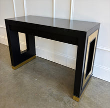 Load image into Gallery viewer, The Ashleigh Console by Theodore Alexander pairs a streamlined silhouette with dramatic decorative details. Professionally lacquered in black, it features substantial rectangular supports accented by textured gold panels, inset antiqued mirror, and brass-capped feet. Two discreet drawers provide concealed storage. Good Bones Furniture
