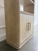 Load image into Gallery viewer, The Horizon Etagere by Theodore Alexander combines generous display space with a light, contemporary silhouette. Crafted from oak in a pale Day finish, its softly rounded frame and finely reeded details introduce texture while preserving the collection’s clean lines. Multiple open shelves provide ample room for books, art, and collected objects, while the two-door cabinet below offers convenient concealed storage. Good Bones Furniture
