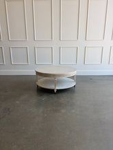 Load image into Gallery viewer, The Lucienne Cocktail Table by Theodore Alexander combines classical ornament with a light, graceful silhouette. Crafted from Khaya wood and richly figured Khaya swirl veneer, its round top rests above a fluted frieze accented with carved leaf motifs. Slender fluted legs connect the upper surface to a wide circular lower shelf.  Washed Blanchette finish. Good Bones Furniture
