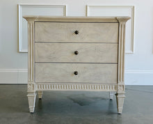 Load image into Gallery viewer, The Valette Nightstand by Theodore Alexander combines generous bedside storage with graceful architectural detail. Crafted from Khaya wood and richly figured Khaya swirl veneer, its three spacious drawers are accented with sculptural floral metal pulls. Fluted corner posts frame the case, while carved leaf motifs, a fluted apron, and tapered legs introduce classical character without overwhelming the clean silhouette. Good Bones Furniture
