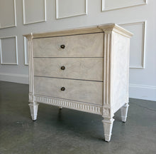 Load image into Gallery viewer, The Valette Nightstand by Theodore Alexander combines generous bedside storage with graceful architectural detail. Crafted from Khaya wood and richly figured Khaya swirl veneer, its three spacious drawers are accented with sculptural floral metal pulls. Fluted corner posts frame the case, while carved leaf motifs, a fluted apron, and tapered legs introduce classical character without overwhelming the clean silhouette. Good Bones Furniture
