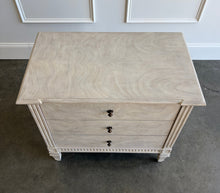 Load image into Gallery viewer, The Valette Nightstand by Theodore Alexander combines generous bedside storage with graceful architectural detail. Crafted from Khaya wood and richly figured Khaya swirl veneer, its three spacious drawers are accented with sculptural floral metal pulls. Fluted corner posts frame the case, while carved leaf motifs, a fluted apron, and tapered legs introduce classical character without overwhelming the clean silhouette. Good Bones Furniture
