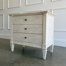 Load image into Gallery viewer, The Valette Nightstand by Theodore Alexander combines generous bedside storage with graceful architectural detail. Crafted from Khaya wood and richly figured Khaya swirl veneer, its three spacious drawers are accented with sculptural floral metal pulls. Fluted corner posts frame the case, while carved leaf motifs, a fluted apron, and tapered legs introduce classical character without overwhelming the clean silhouette. Good Bones Furniture
