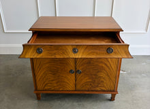 Load image into Gallery viewer, The Valencia Bedside Chest combines elegant architectural detailing with beautifully figured wood grain. Crafted from mahogany solids with richly patterned cerejeira veneers, it features two cabinet doors that open to reveal a spacious interior with one adjustable shelf. molded top, graceful concave frieze, decorative metal hardware, and fluted tapered feet. Good Bones Furniture
