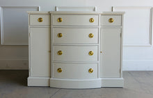 Load image into Gallery viewer, This vintage buffet combines generous storage with a compact, beautifully balanced silhouette. Professionally lacquered in a warm Custom Cream, it features a subtly bowed center with four spacious drawers, two smaller side drawers, and flanking cabinets with interior shelves. New brass Hepplewhite hardware. Good Bones Furniture
