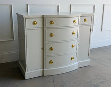 Load image into Gallery viewer, This vintage buffet combines generous storage with a compact, beautifully balanced silhouette. Professionally lacquered in a warm Custom Cream, it features a subtly bowed center with four spacious drawers, two smaller side drawers, and flanking cabinets with interior shelves. New brass Hepplewhite hardware. Good Bones Furniture
