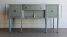 Load image into Gallery viewer, This vintage sideboard pairs a historic Hepplewhite-inspired silhouette with an unexpected modern color. Its gently bowed front, symmetrical layout, slender tapered legs, and balanced proportions reflect classic Federal design. Professionally lacquered in Sherwin-Williams Unusual Gray with new solid brass oval bail pulls with ornate neoclassical backplates. Good Bones Furniture
