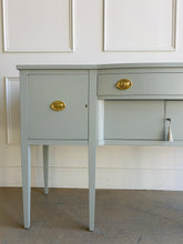 Load image into Gallery viewer, This vintage sideboard pairs a historic Hepplewhite-inspired silhouette with an unexpected modern color. Its gently bowed front, symmetrical layout, slender tapered legs, and balanced proportions reflect classic Federal design. Professionally lacquered in Sherwin-Williams Unusual Gray with new solid brass oval bail pulls with ornate neoclassical backplates. Good Bones Furniture
