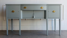 Load image into Gallery viewer, This vintage sideboard pairs a historic Hepplewhite-inspired silhouette with an unexpected modern color. Its gently bowed front, symmetrical layout, slender tapered legs, and balanced proportions reflect classic Federal design. Professionally lacquered in Sherwin-Williams Unusual Gray with new solid brass oval bail pulls with ornate neoclassical backplates. Good Bones Furniture
