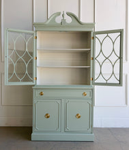 Load image into Gallery viewer, This vintage china cabinet blends classic form with a fresh, tailored finish. Professionally lacquered in Benjamin Moore Saybrook Sage on the exterior and paired with a creamy white interior, it offers a soft contrast that brightens the display space and gives the piece a custom, updated feel. Arched fretwork glass doors and a sculptural pediment. Good Bones Furniture
