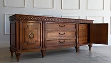 Load image into Gallery viewer, This vintage Mediterranean/ Spanish Revival credenza dresser has the scale, craftsmanship, and presence that make 1970s American case goods so collectible. The design features a carved scrolling frieze, burl veneer door panels, architectural legs, and original brass ring hardware with geometric backplates. Good Bones Furniture

