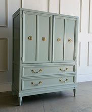 Load image into Gallery viewer, This vintage Thomasville armoire professionally lacquered in a custom muted green with soft blue-gray undertones, while the interior is finished in French Roast. Four paneled doors open to reveal divided upper compartments and two pull-out tray drawers. Two spacious lower drawers offer additional concealed storage. Good Bones Furniture
