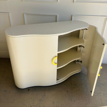 Load image into Gallery viewer, Three-quarter view of the Highland House Jules Cabinet with one door open, showcasing its architectural curves, interior shelving, and brass hardware against a creamy white lacquered finish.
